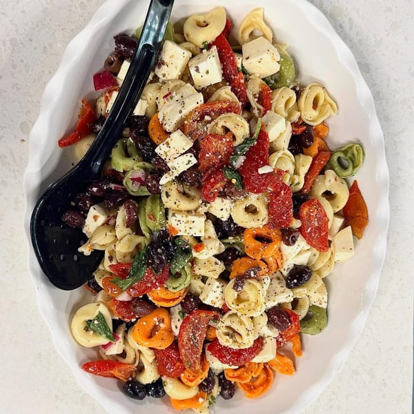 A white platter filled with colorful tortellini pasta salad, feta, sun-dried tomatoes, and olives.